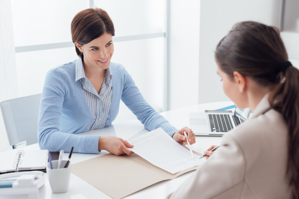 A woman at a office desk showing another woman a pad of paper